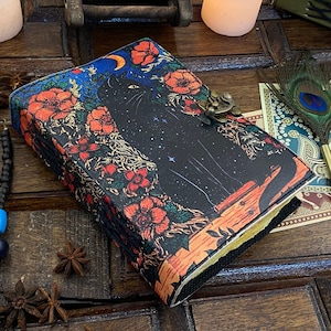May include: A black leather journal with a colourful floral design featuring a black cat silhouette against a starry night sky. The journal has a brass clasp closure.