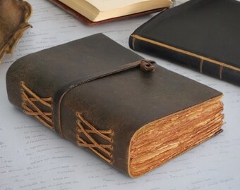 May include: A brown leather-bound journal with a distressed look. The journal has a strap closure and is tied with a brown leather cord. The journal is lying on a white surface.
