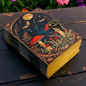 K&ouml;nnte beinhalten: Ein dekoratives Notizbuch mit einem himmlischen und Pilz-Design. Das Cover zeigt einen Nachthimmel mit Vollmond, Schmetterlingen und roten Pilzen. Die Seiten haben einen goldenen Rand, und das Notizbuch ist mit einem Metallverschluss gesichert.