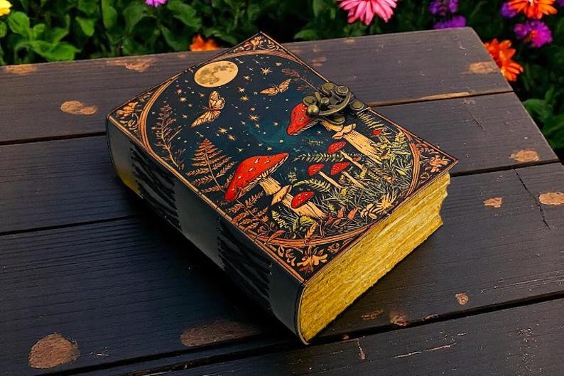 K&ouml;nnte beinhalten: Ein dekoratives Notizbuch mit einem himmlischen und Pilz-Design. Das Cover zeigt einen Nachthimmel mit Vollmond, Schmetterlingen und roten Pilzen. Die Seiten haben einen goldenen Rand, und das Notizbuch ist mit einem Metallverschluss gesichert.