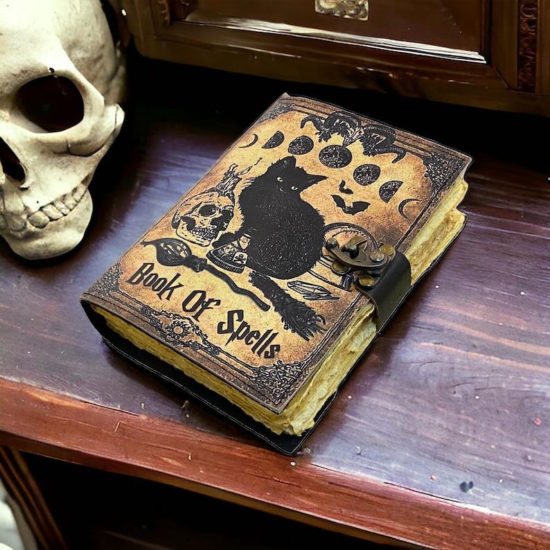 Puede incluir: Un diario encuadernado en cuero marr&oacute;n con un gato negro, una calavera y otras im&aacute;genes de brujer&iacute;a. La portada dice "Book of Spells".