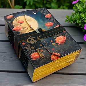 Könnte beinhalten: Ein dekoratives Notizbuch mit floralem und himmlischem Design. Das Cover zeigt einen Nachthimmel mit Vollmond und pfirsichfarbenen Blumen. Ein schwarzer Riemen und ein Schlüsselverschluss sichern das Notizbuch. Die Seitenränder sind goldfarben.