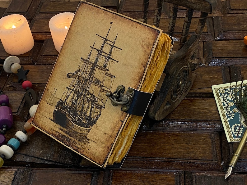 Pode incluir: Um di&aacute;rio de estilo vintage encadernado em couro com uma ilustra&ccedil;&atilde;o desenhada &agrave; m&atilde;o de um navio &agrave; vela na capa. O di&aacute;rio tem um fecho de lat&atilde;o e uma apar&ecirc;ncia amarelada e envelhecida.