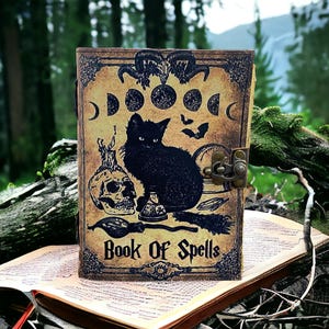 Puede incluir: Un diario de cuero marr&oacute;n con un gato negro, una calavera y otras im&aacute;genes de brujer&iacute;a. La portada dice "Book of Spells".