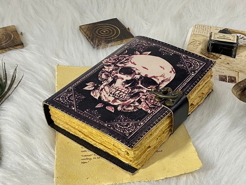 K&ouml;nnte beinhalten: Ein schwarzes, ledergebundenes Tagebuch mit einem Totenkopf- und Rosenmotiv. Das Tagebuch hat einen Messingverschluss und einen schwarzen Lederriemen.