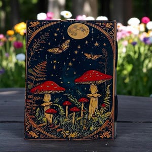 K&ouml;nnte beinhalten: Ein dekoratives Notizbuch mit einem skurrilen Design mit roten Pilzen, Schmetterlingen und einem Vollmond vor einem dunkelblauen Sternenhimmel. Das Notizbuch hat einen verzierten Goldrand und befindet sich vor einem verschwommenen Hintergrund im Freien.