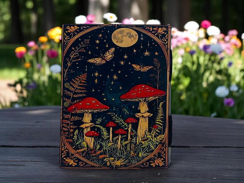 K&ouml;nnte beinhalten: Ein dekoratives Notizbuch mit einem skurrilen Design mit roten Pilzen, Schmetterlingen und einem Vollmond vor einem dunkelblauen Sternenhimmel. Das Notizbuch hat einen verzierten Goldrand und befindet sich vor einem verschwommenen Hintergrund im Freien.