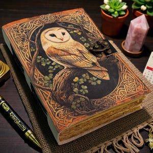 May include: An ornate journal with an owl design on the cover, framed by Celtic knotwork. The journal has a metal clasp and aged pages. A lit candle, pen, and a 2025 calendar are also visible.