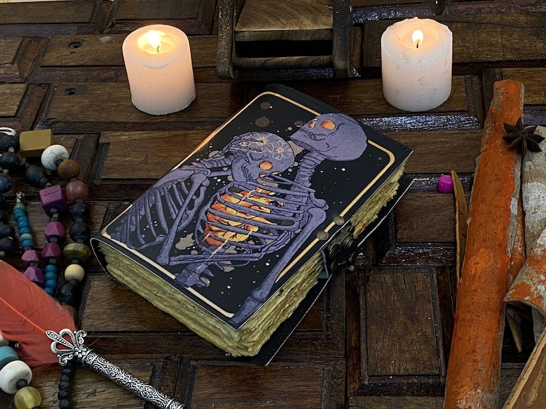 May include: A black leather-bound journal with a detailed illustration of two skeletons embracing. The skeletons are purple and white, and the background is a dark blue with white stars. The journal is closed and has a brass clasp.