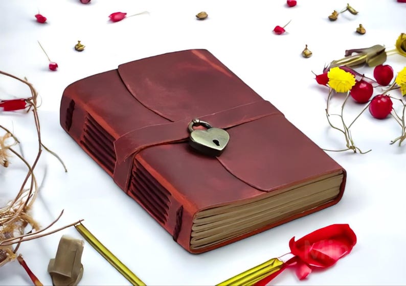 May include: A burgundy leather-bound journal with a heart-shaped lock and a decorative strap. The journal is closed and lying on a white surface.