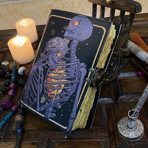 May include: A black leather-bound journal with a colorful illustration of two skeletons embracing. The journal has a distressed, aged look and is closed. The journal is sitting on a wooden surface with candles and other decorative items.