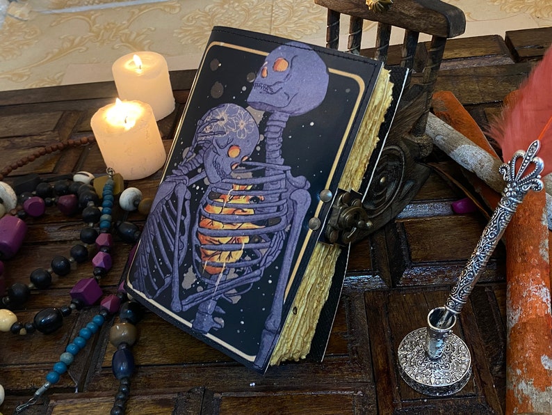 May include: A black leather-bound journal with a colorful illustration of two skeletons embracing. The journal has a distressed, aged look and is closed. The journal is sitting on a wooden surface with candles and other decorative items.