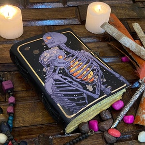 May include: A black leather journal with a galaxy-themed cover featuring two skeletons embracing. The skeletons are purple and have a glowing orange heart. The journal is surrounded by candles, feathers, and beads.