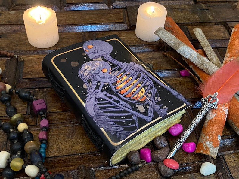 May include: A black leather journal with a galaxy-themed cover featuring two skeletons embracing. The skeletons are purple and have a glowing orange heart. The journal is surrounded by candles, feathers, and beads.