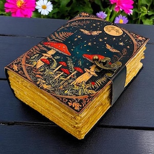 K&ouml;nnte beinhalten: Ein ledergebundenes Notizbuch mit einem Fantasy-Cover mit roten Pilzen, Schmetterlingen und einem Mond vor einem dunkelblauen Hintergrund. Das Notizbuch hat gealterte, gelbliche Seiten und einen Metallverschluss mit einem schwarzen Lederriemen.