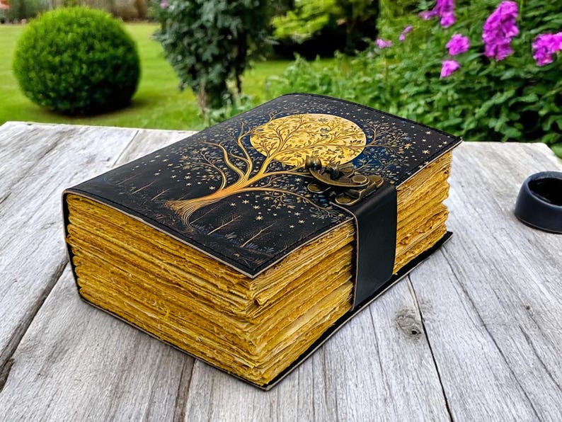 May include: A black leather-bound journal with a gold tree and moon design. The journal has a clasp closure and is made of aged paper.