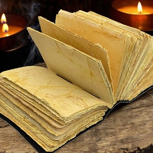 May include: An open book with aged, yellowed pages and a black binding rests on a wooden surface. Two lit candles in copper holders and wisps of smoke create a mystical atmosphere in the background.