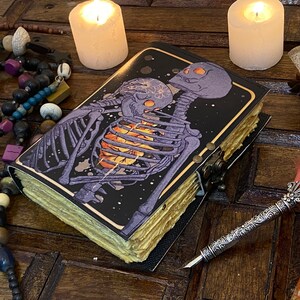 May include: A black leather-bound journal with a gold trim and a colorful illustration of two skeletons embracing. The journal is closed and lying on a wooden surface with a quill pen and other decorative items.