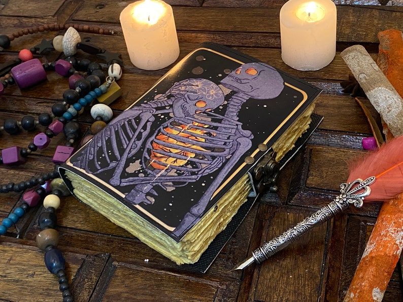 May include: A black leather-bound journal with a gold trim and a colorful illustration of two skeletons embracing. The journal is closed and lying on a wooden surface with a quill pen and other decorative items.