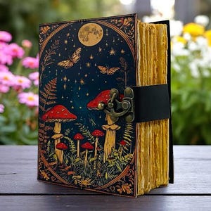 K&ouml;nnte beinhalten: Ein dekoratives Notizbuch mit Fantasy-Motiv. Das Cover zeigt eine Nachtszene mit Pilzen, Schmetterlingen und einem Vollmond. Die Seiten sind gelblich gealtert, und das Notizbuch wird mit einem schwarzen Lederriemen und einem Metallverschluss gesichert.