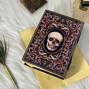 K&ouml;nnte beinhalten: Ein schwarzes, ledergebundenes Tagebuch mit einem Totenkopf- und Blumendesign. Das Tagebuch hat einen abgenutzten Look und ist mit goldenen Akzenten verziert.