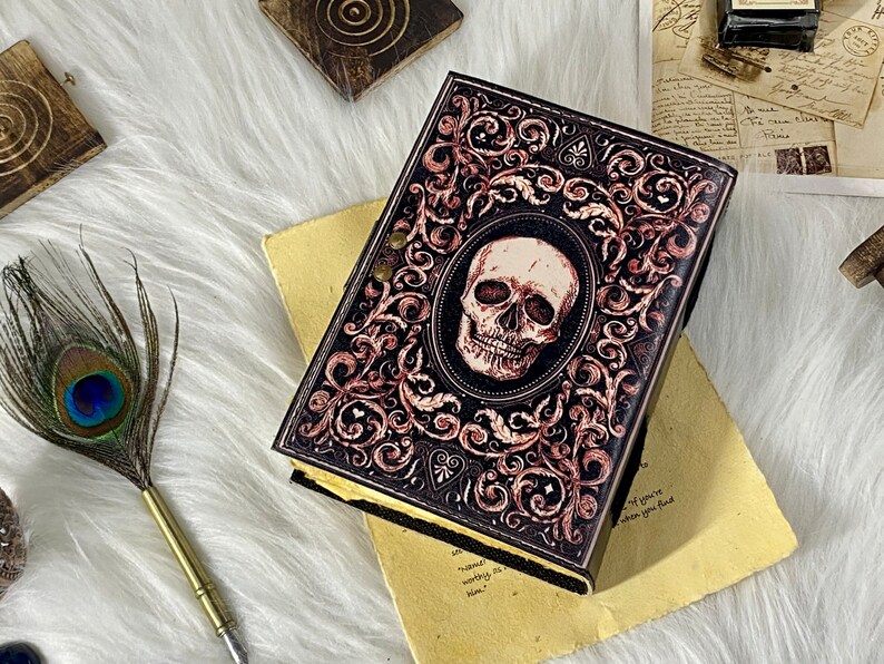 K&ouml;nnte beinhalten: Ein schwarzes, ledergebundenes Tagebuch mit einem Totenkopf- und Blumendesign. Das Tagebuch hat einen abgenutzten Look und ist mit goldenen Akzenten verziert.