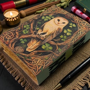 May include: A detailed wooden box featuring a barn owl illustration, Celtic knotwork, and shamrocks. The box sits on a burlap cloth, with candles and a pen. The box is likely used for storage.