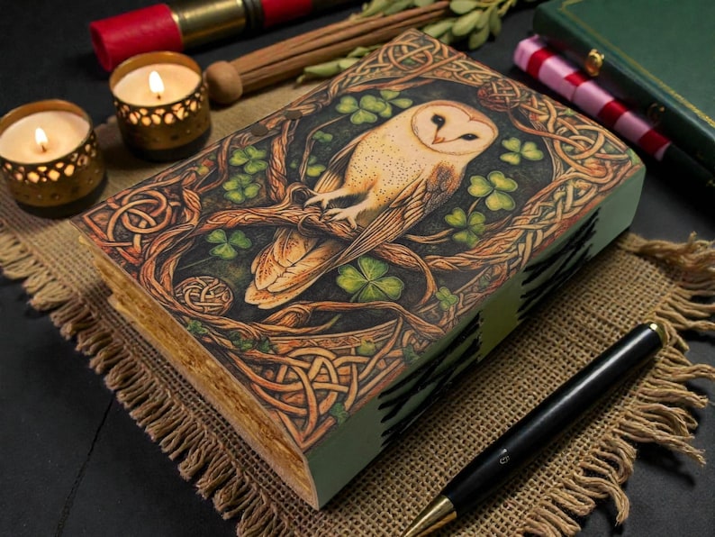 May include: A detailed wooden box featuring a barn owl illustration, Celtic knotwork, and shamrocks. The box sits on a burlap cloth, with candles and a pen. The box is likely used for storage.