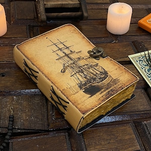 Pode incluir: Um di&aacute;rio encadernado em couro com uma ilustra&ccedil;&atilde;o de estilo vintage de um navio &agrave; vela na capa. O di&aacute;rio tem um fecho de lat&atilde;o e costuras decorativas ao redor das bordas.