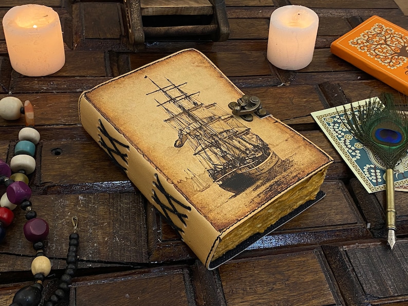 Pode incluir: Um di&aacute;rio encadernado em couro com uma ilustra&ccedil;&atilde;o de estilo vintage de um navio &agrave; vela na capa. O di&aacute;rio tem um fecho de lat&atilde;o e costuras decorativas ao redor das bordas.