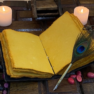 May include: An open journal with blank pages, a peacock feather quill pen, and two white candles on a wooden surface.
