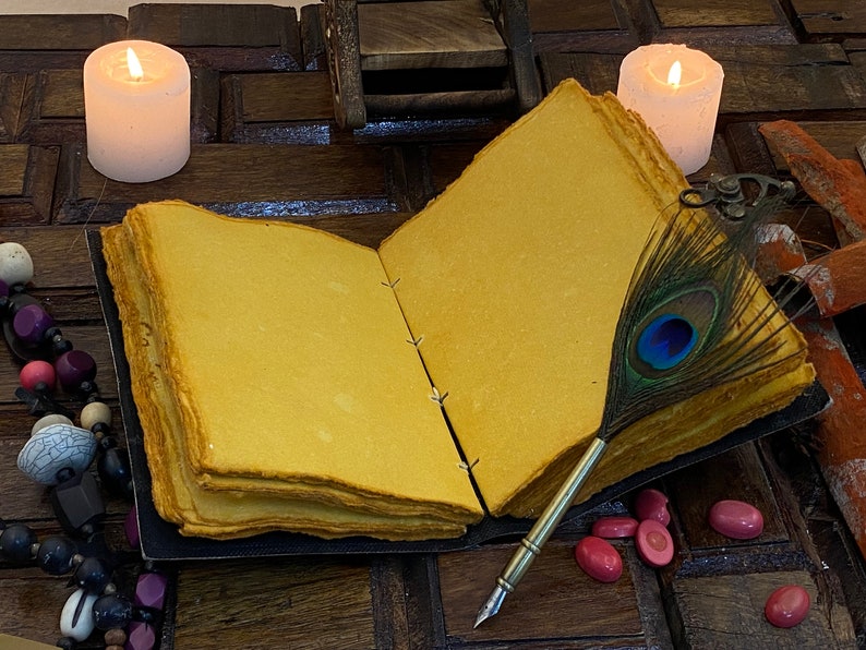 May include: An open journal with blank pages, a peacock feather quill pen, and two white candles on a wooden surface.