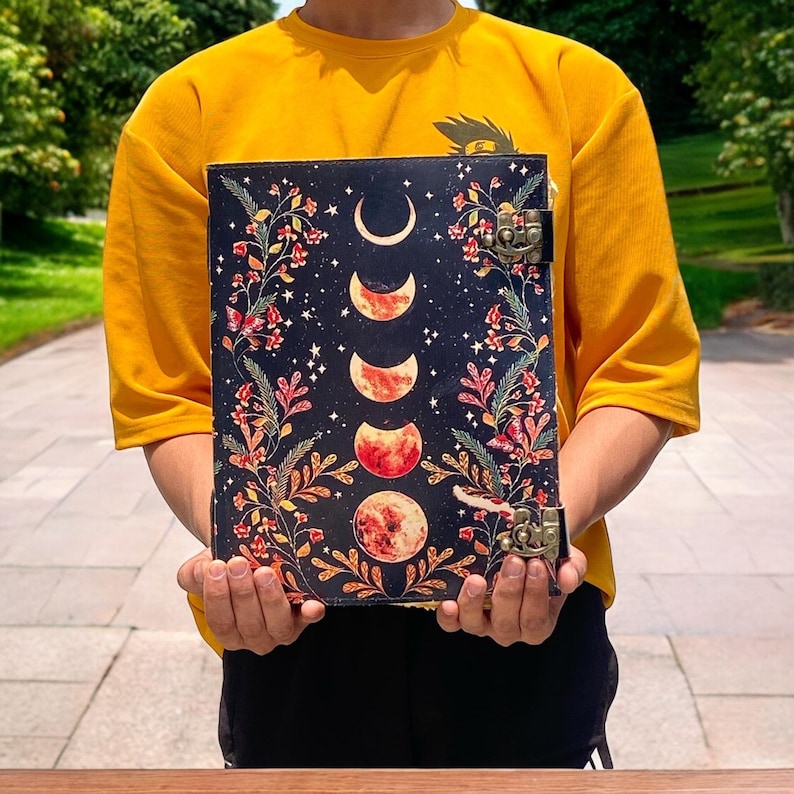 May include: A black leather journal with a floral design and a moon phase pattern. The journal has two brass clasps and is held by a person wearing a yellow shirt.