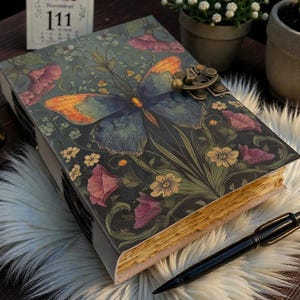 May include: A decorative journal with a butterfly and floral design on the cover. The journal has a metal clasp and aged-looking pages. A black pen rests beside the journal on a fluffy white surface.