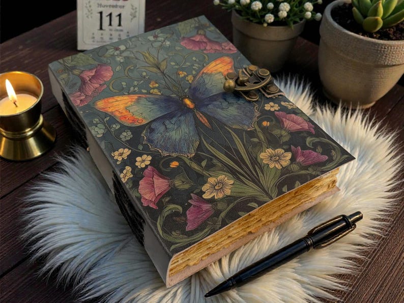 May include: A decorative journal with a butterfly and floral design on the cover. The journal has a metal clasp and aged-looking pages. A black pen rests beside the journal on a fluffy white surface.