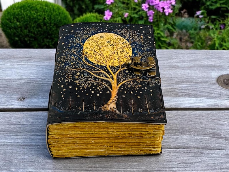 May include: A black leather journal with a gold tree and moon design. The journal has a brass lock and clasp. The pages are yellowed and have a rustic look.