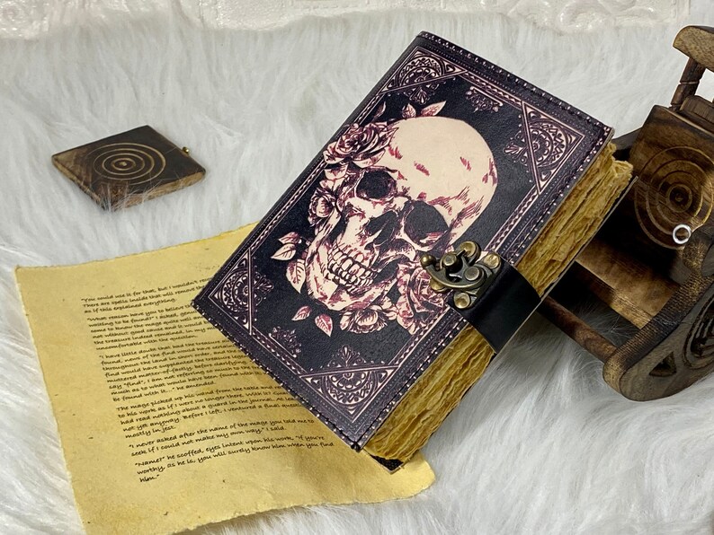 K&ouml;nnte beinhalten: Ein schwarzes Lederjournal mit einem Totenkopf- und Rosenmotiv. Das Journal hat einen Messingverschluss und einen braunen Lederriemen. Das Journal ist geschlossen und liegt auf einer wei&szlig;en Oberfl&auml;che.