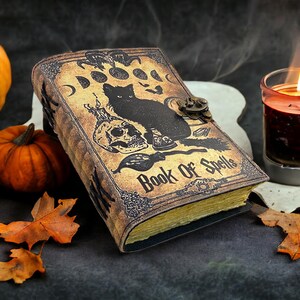 Puede incluir: Un diario encuadernado en cuero con un dise&ntilde;o vintage que presenta un gato negro, una calavera y las palabras "Libro de Hechizos". El diario est&aacute; rodeado de calabazas y velas, creando un ambiente de Halloween espeluznante.