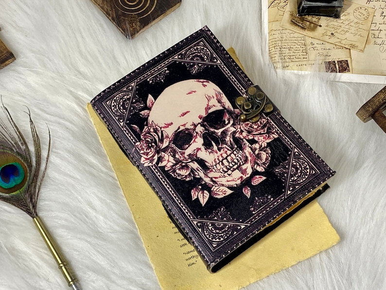 K&ouml;nnte beinhalten: Ein schwarzes Lederjournal mit einem Totenkopf- und Rosenmotiv. Das Journal hat einen Messingverschluss und ist geschlossen.