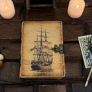 Pode incluir: Um di&aacute;rio de couro de estilo vintage com uma ilustra&ccedil;&atilde;o detalhada de um navio &agrave; vela na capa. O di&aacute;rio tem um fecho de metal e um aspeto desgastado e envelhecido.