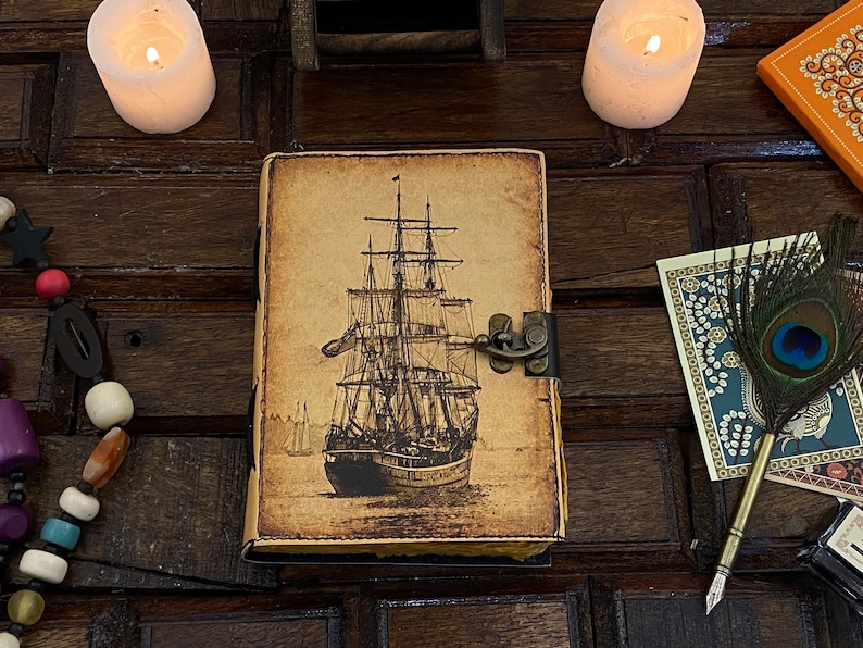 Pode incluir: Um di&aacute;rio de couro de estilo vintage com uma ilustra&ccedil;&atilde;o detalhada de um navio &agrave; vela na capa. O di&aacute;rio tem um fecho de metal e um aspeto desgastado e envelhecido.