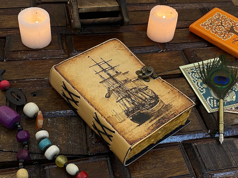 Pode incluir: Um di&aacute;rio encadernado em couro com uma imagem de estilo vintage de um navio &agrave; vela na capa. O di&aacute;rio tem uma capa de couro marrom com costuras decorativas e um fecho de lat&atilde;o.
