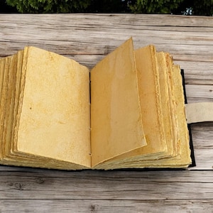 K&ouml;nnte beinhalten: Ein offenes Journal im antiken Stil mit gealterten, vergilbten Seiten. Das Buch hat einen schwarzen Einband und einen Lederriemenverschluss mit Metallverschluss. Die Seiten sind dick und strukturiert. Das Journal liegt auf einer Holzoberfl&auml;che.