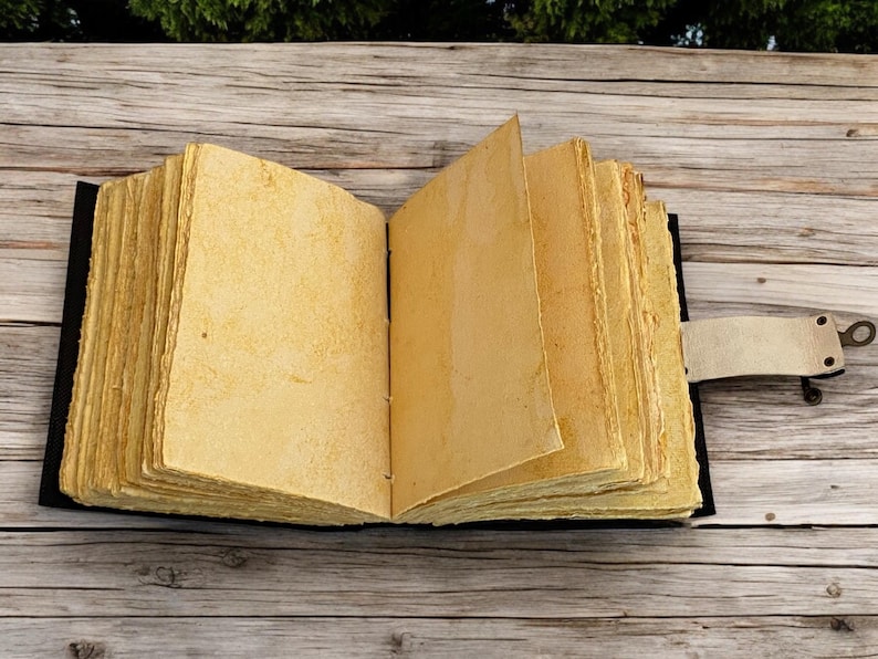 K&ouml;nnte beinhalten: Ein offenes Journal im antiken Stil mit gealterten, vergilbten Seiten. Das Buch hat einen schwarzen Einband und einen Lederriemenverschluss mit Metallverschluss. Die Seiten sind dick und strukturiert. Das Journal liegt auf einer Holzoberfl&auml;che.