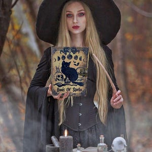 Puede incluir: Una mujer vestida de bruja sostiene un libro con un gato negro y las palabras "Book of Spells" en la portada. Est&aacute; de pie en un entorno de bosque con una mesa frente a ella, que tiene una vela, un mortero y un pil&oacute;n, y otros art&iacute;culos de brujer&iacute;a.