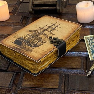 Pode incluir: Um di&aacute;rio de estilo vintage encadernado em couro com uma ilustra&ccedil;&atilde;o de um navio na capa. O di&aacute;rio tem um fecho de metal e uma correia de couro preto. O di&aacute;rio est&aacute; sobre uma superf&iacute;cie de madeira com velas e outros itens decorativos.