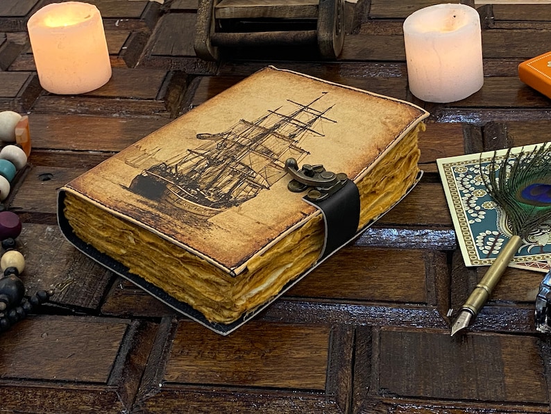 Pode incluir: Um di&aacute;rio de estilo vintage encadernado em couro com uma ilustra&ccedil;&atilde;o de um navio na capa. O di&aacute;rio tem um fecho de metal e uma correia de couro preto. O di&aacute;rio est&aacute; sobre uma superf&iacute;cie de madeira com velas e outros itens decorativos.