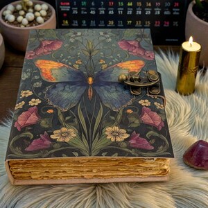 May include: A decorative journal with a butterfly design on the cover. The butterfly has blue and orange wings, set against a dark background with floral accents. The journal has a metal clasp and aged-looking pages.