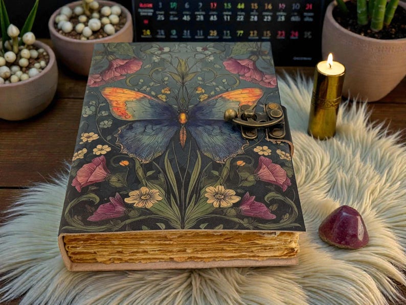 May include: A decorative journal with a butterfly design on the cover. The butterfly has blue and orange wings, set against a dark background with floral accents. The journal has a metal clasp and aged-looking pages.