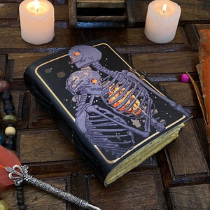 May include: A black leather-bound journal with a dark purple and gold illustration of two skeletons embracing. The skeletons are on a black background with a starry pattern. The journal is on a wooden surface with candles and other decorative items.